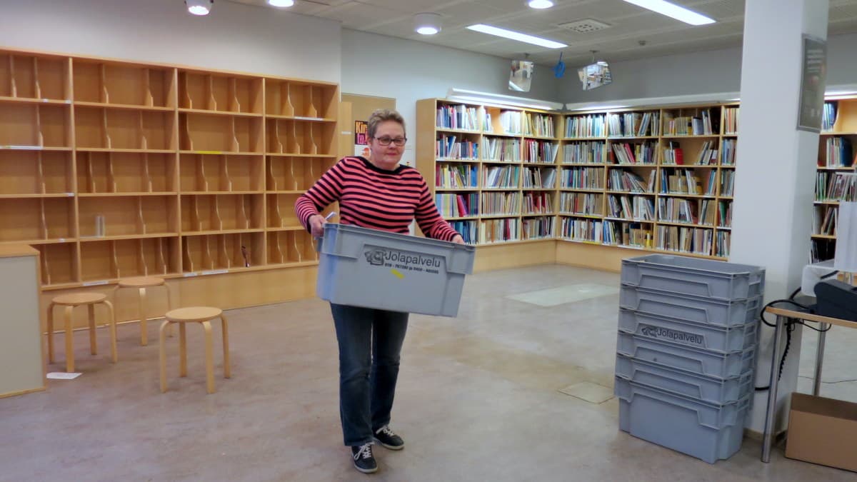 Nainen kantaa painavaa laatikkoa puolityhjässä kirjastohuoneessa