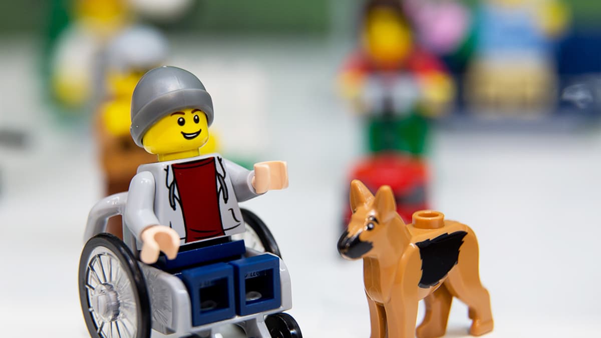 Pyörätuolissa istuva Lego-hahmo avustajakoiran seurassa.