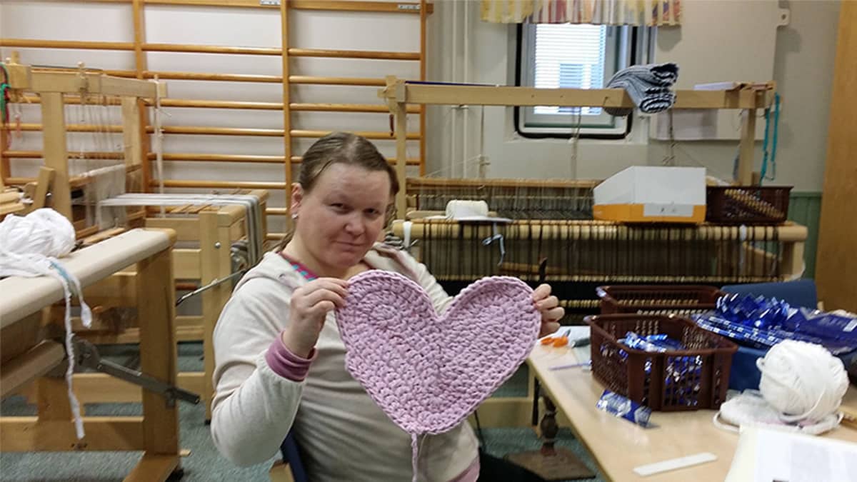 Nina Pyykkönen esittelee virkattua vaaleanpunaista sydäntä työpajassa. Taustalla kangaspuita.