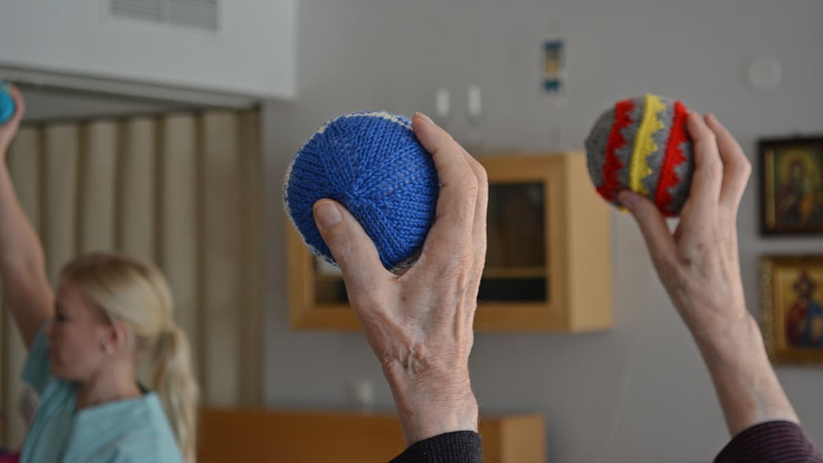 Vanhukset pitelevät neulottuja kuntopalloja ilmassa käsissään. Taustalla fysioterapeutti Hanna Väisänen näyttää mallia. 