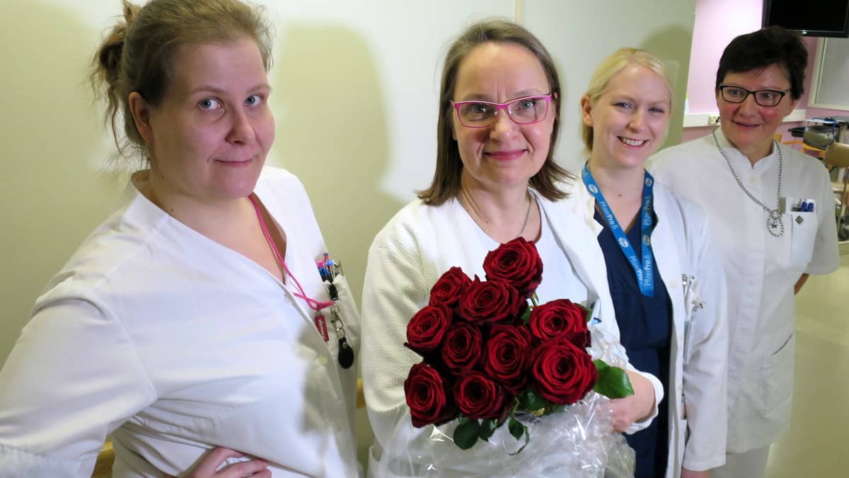 Neljä naista katsoo sairaalan käytävällä kameraan. Heistä keskimmäisellä on iso ruusukimppu.