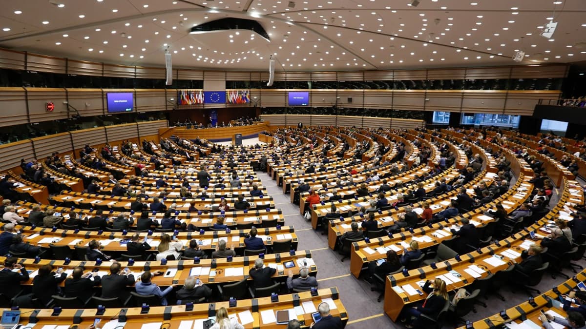 Kuva Euroopan parlamentin istuntosalista, jossa edustajien pöydät levittäytyvät puheenjohtajan korokkeesta. Puheenjohtajan korokkeen yllä on EU:n lippu ja jäsenvaltioiden liput.