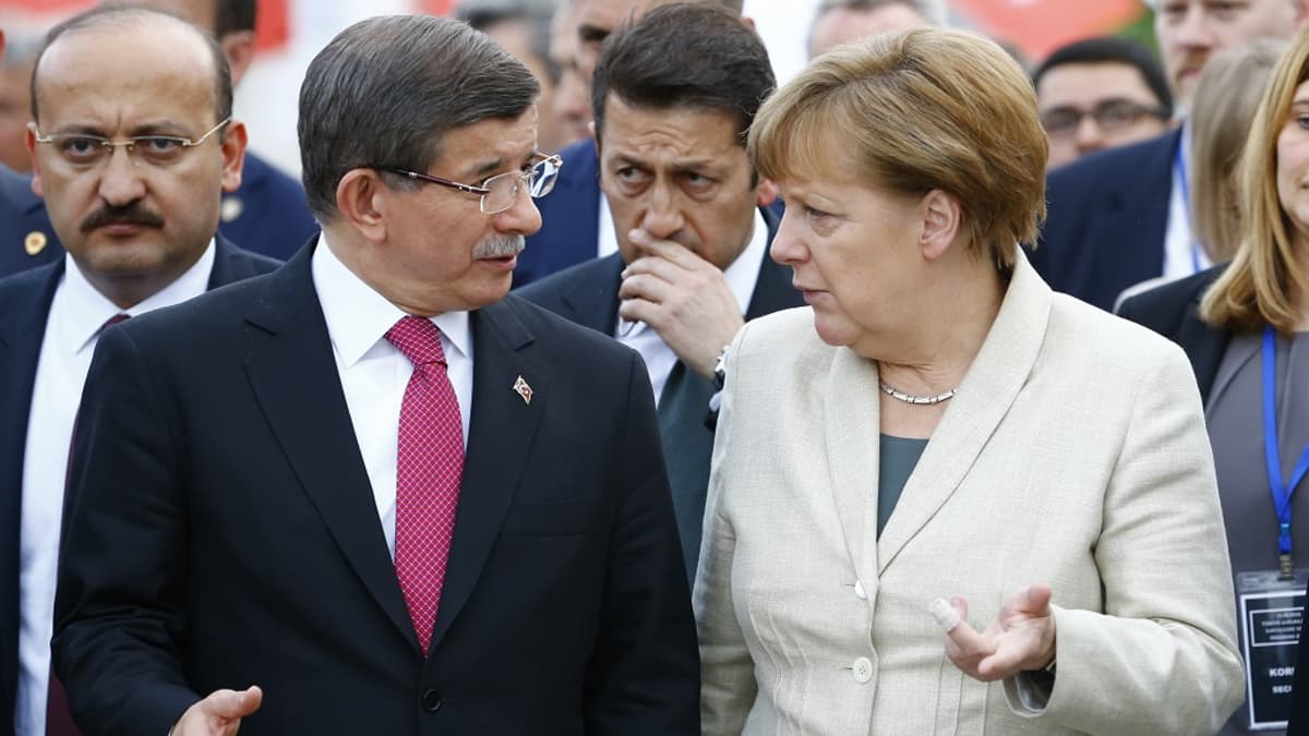 Ahmet Davutoğlu ja Angela Merkel keskustelevat. Turkin pääministerillä on tumma puku ja punainen ruudukas kravatti, Saksan liittokanslerilla vaalea bleiseri, vihreä paita. Taustalla näkyy virkamiehiä.