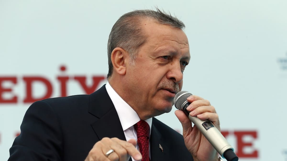 Erdoğan puhuu vasemmassa kädessään olevaan mikrofoniin ja osoittaa oikean kätensä etusormella alaspäin. Hänellä on tumma puku, punainen ruudullinen kravatti. 