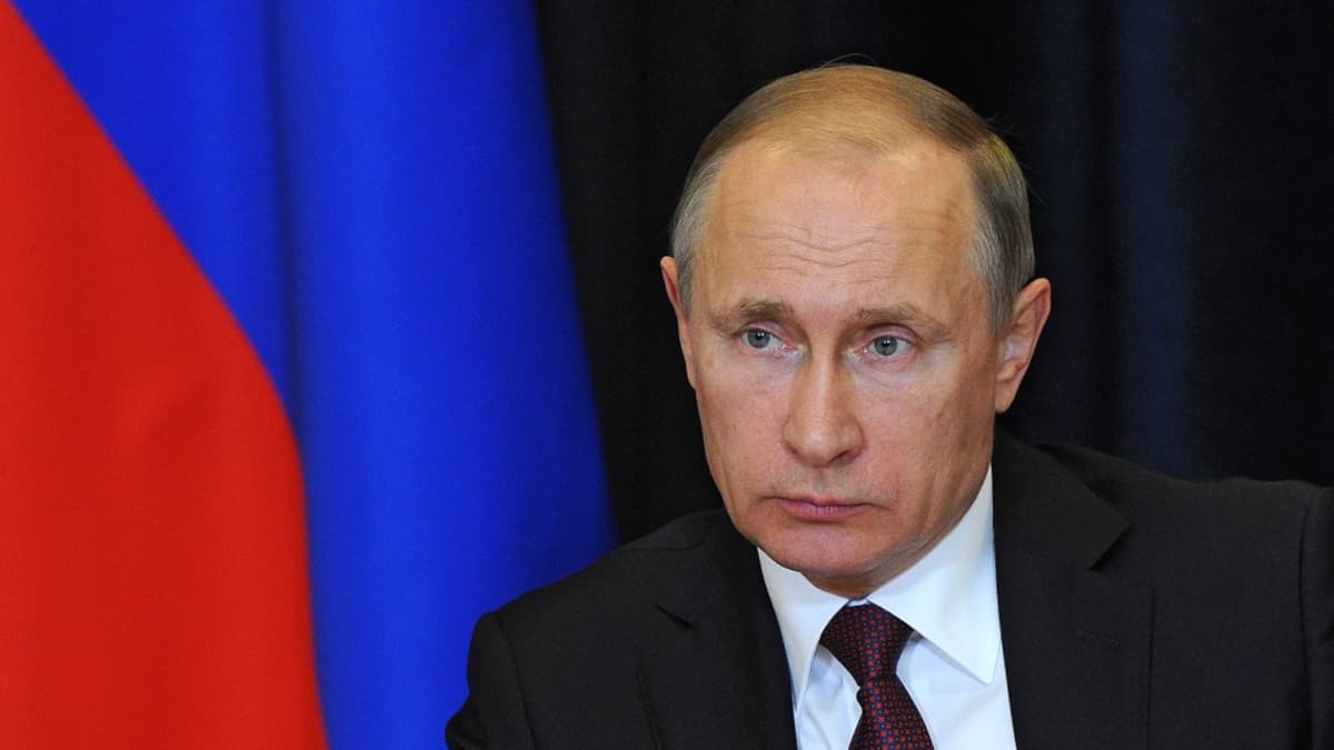 Putin katsoo vakavana kuvan vasenta reunaa kohti. Hänellä on tumma puku, tummanpunainen kravatti, jossa on sinistä kuviota. Taustalla näkyy Venäjän lippu.