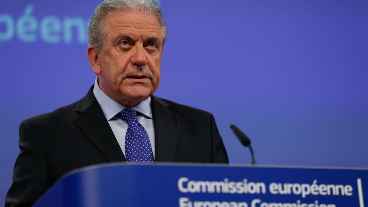 Komissaari seisoo puhujakorokkeen takana tummassa puvussa, sininen pilkullinen kravatti kaulassaan. Hänen edessään on mikrofoni. Tausta on EU:n sininen.