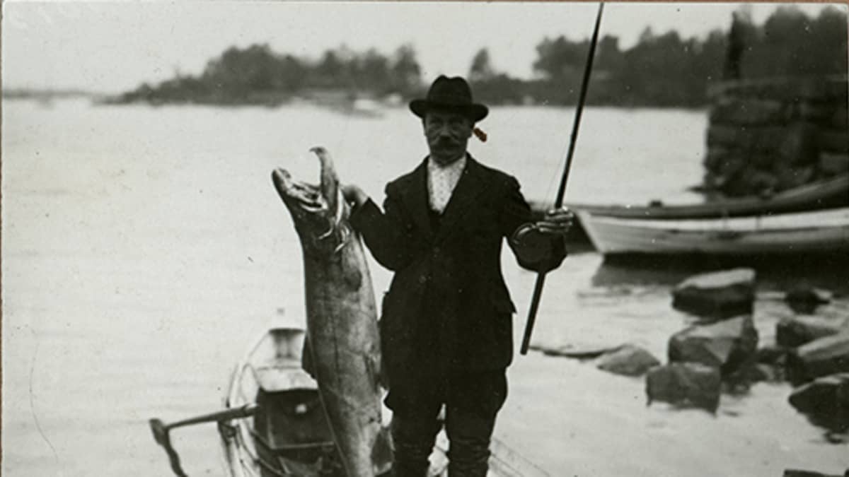 Vanhassa, mustavalkoisessa kuvassa mies seisoo veneessä lohisaaliinsa kanssa.