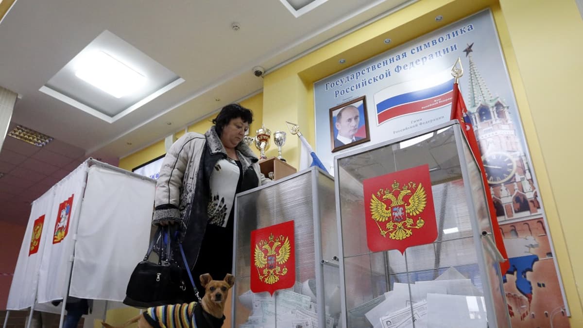 Koiraa taluttava nainen laittaa vaalilipuketta uurnaan. Takana näkyy äänestyskoppeja ja seinällä Putinin kuva.
