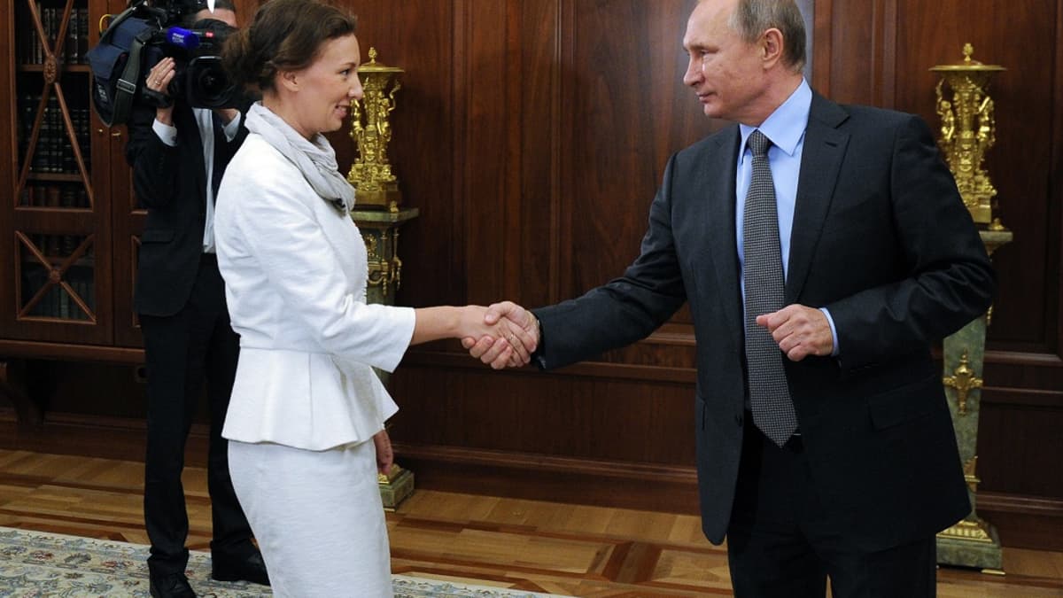 Anna Kuznetsova ja Vladimir Putin kättelevät Putinin työhuoneessa, jonka seinät ovat tummaa puupaneelia. Molemmat hymyilevät. Kuznetsovalla on valkoinen asu, Putinilla tumma puku, vaaleansininen kauluspaita ja kravatti. Taustalla kirjahyllyn edessä näkyy television kameramies, joka kuvaa tilannetta.