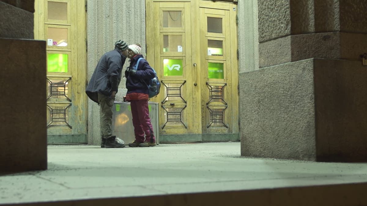 Asunnoton pariskunta vaihtaa suudelman Helsingin rautatieaseman edessä. 