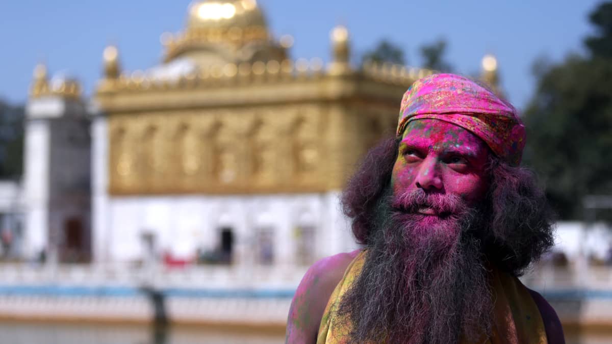 Turbaanipäinen pitkätukkainen mies seisoo temppelin pihalla. Hänet on värjättyeri väreillä.