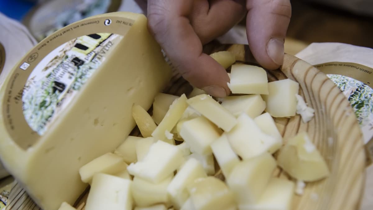 Miehen käsi poimii juustonpalaa vadilta, johon on kuutioitu puolikas juustokiekosta. 