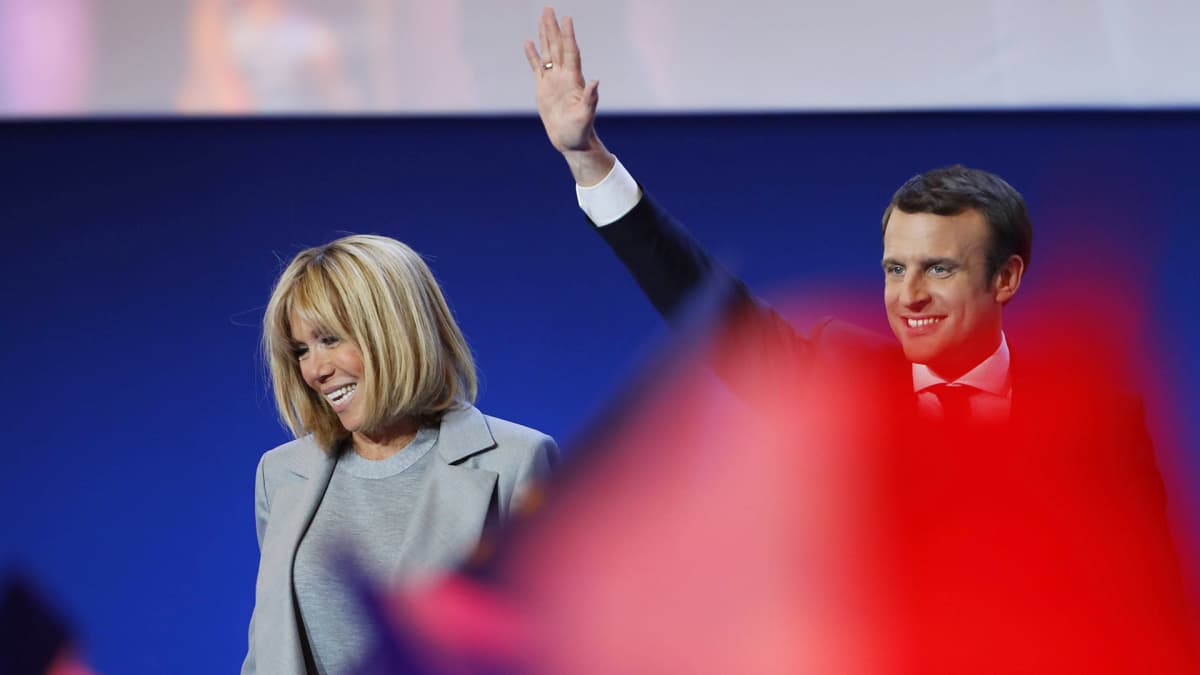 Brigitte Trogneux ja Emmanuel Macron seisovat esiintymislavalla. Macron heiluttaa kättään. Etualalla joku heiluttaa jonkinlaista punaista vaatetta, joka näkyy kuvassa punaisen värin leiskahduksena.