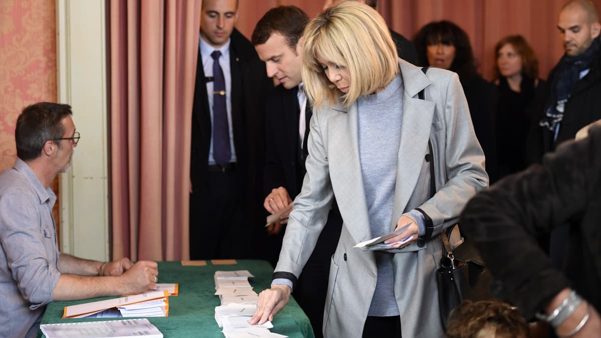 Brigitte Trogneux ja Emmanuel Macron poimivat äänestyksessä tarvittavia lappusia vaalivirkailijan valvomalta pöydältä. Trogneux'lla on vaaleanharmaa päällystakki. Macronilla on tumma puku.