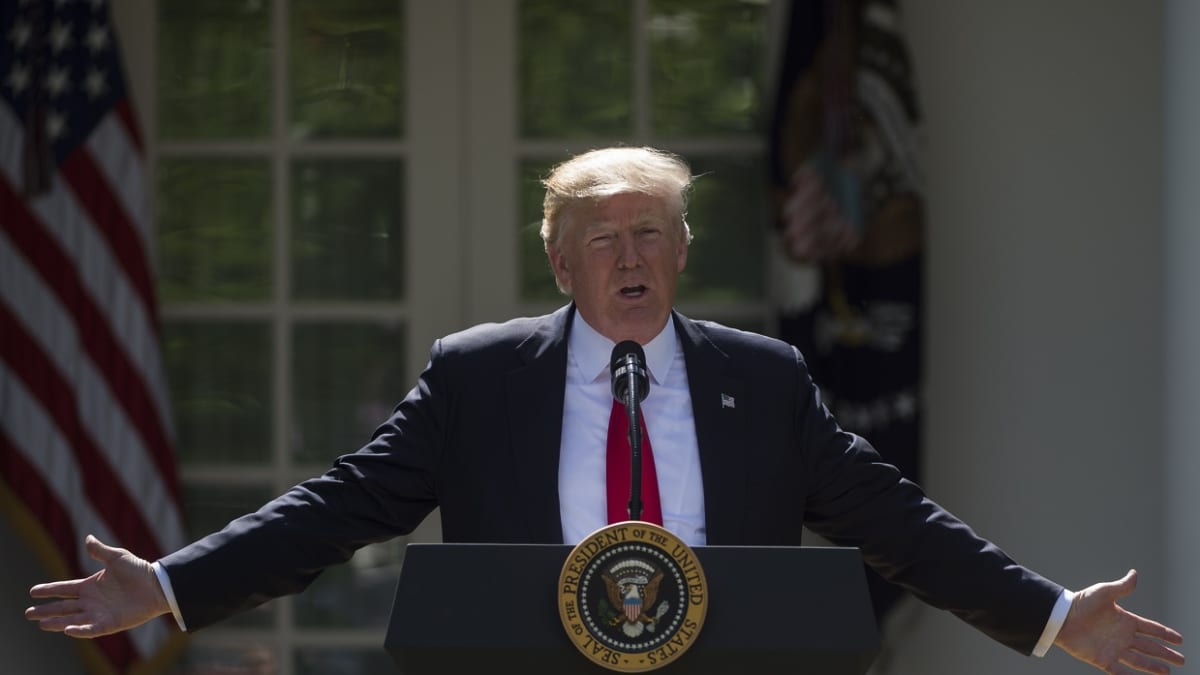 Trump puhujapöntössä valkoisen talon pihalla, levittelee käsiään. 