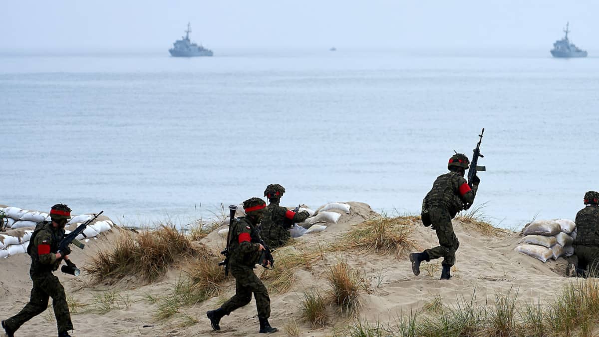 Sotilaat juoksevat hiekassa, taustalla meri jossa kaksi alusta.