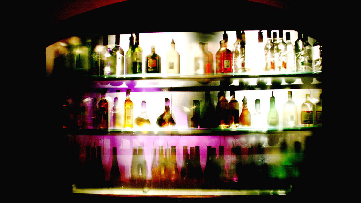Alkoholipulloja baarin kaapissa rivissä, taustalta tulee valoa pullojen läpi.