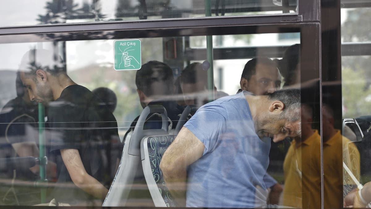 Siviilivaatteissa olevia miehiä näkyy linja-auton ikkunan läpi. Heillä on kädet selän takana.