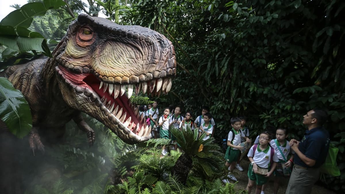 Pensaikosta kurkisteleennollisen kokoinen T. rex -robotti. Puiston opas kertoo siitä koulupukuiselle joukolle alakouluikäisiä lapsia. 