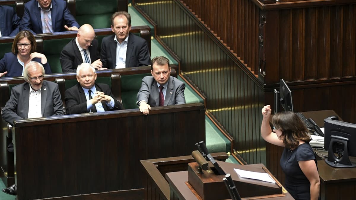 Miehiä tummissa puvuissa ja nainen mustassa mekossa puhujanpöntössä.