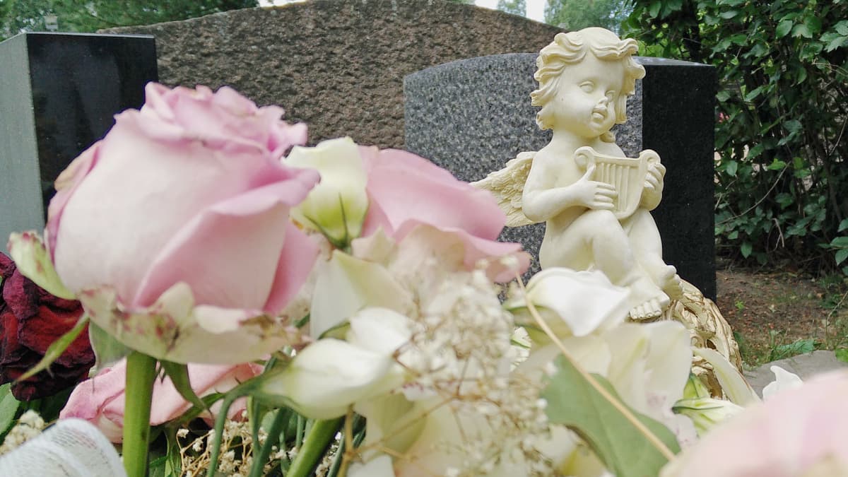 Pieni, valkoinen enkelipatsas haudalla ja etualalla ruusukimppu.