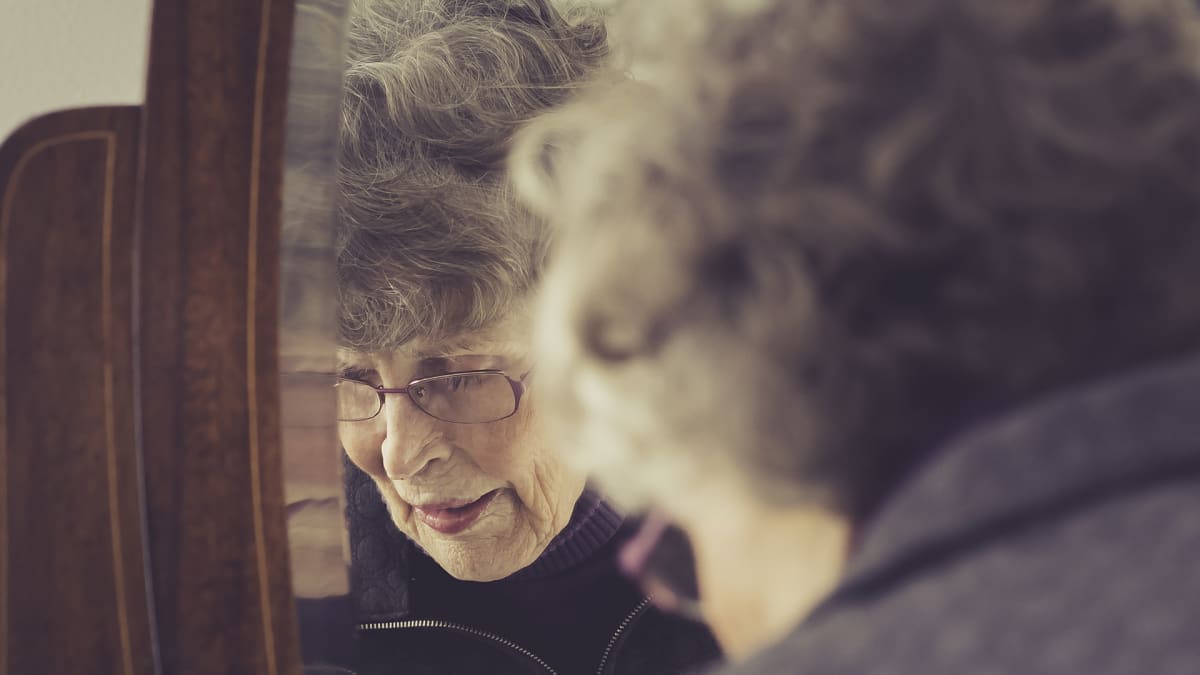Vanhempi nainen siirtelee tavaroita peilipiirongin päällä ja hänen kasvonsa heijastuvat peilin kautta kohti kameraa.