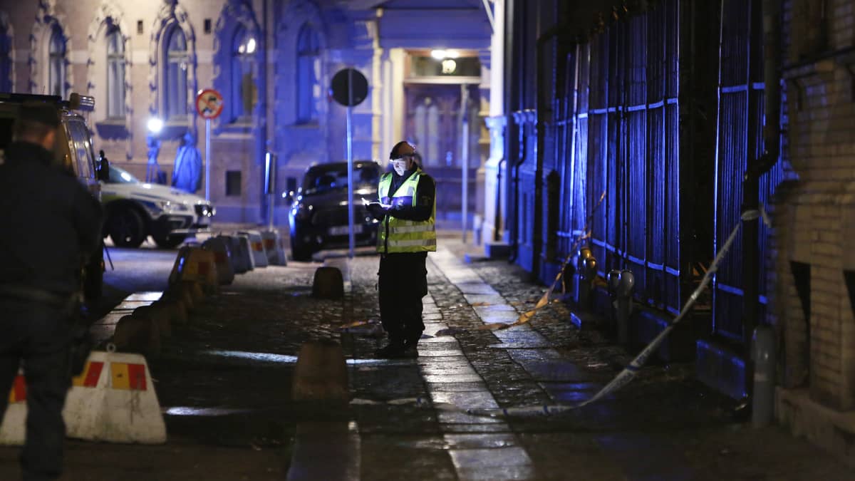 Kuva öiseltä kadulta. Seinästä roikkuu poliisin eristysnauhaa. Kuvan keskellä seisoo poliisimies keltaisissa huomioliiveissä.