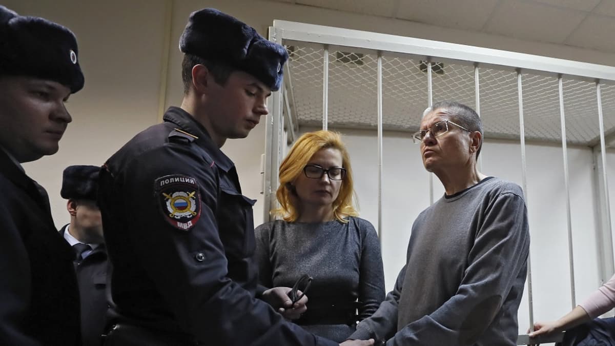 Poliisi laittaa käsirautoja harmaapaitaisen, silmälasipäisen Uljukajevin ranteisiin. Vieressä seisoo Uljukajevin vaimo. Taustalla on häkki, jossa syytetyt istuvan oikeudenkäynnin aikana.
