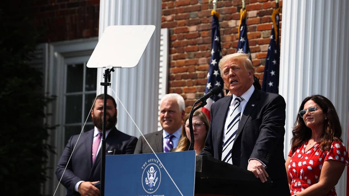 Kuvassa Donald Trump seisoo puhujanpöntön takana. Hänen takanaan on neljä ihmistä. 