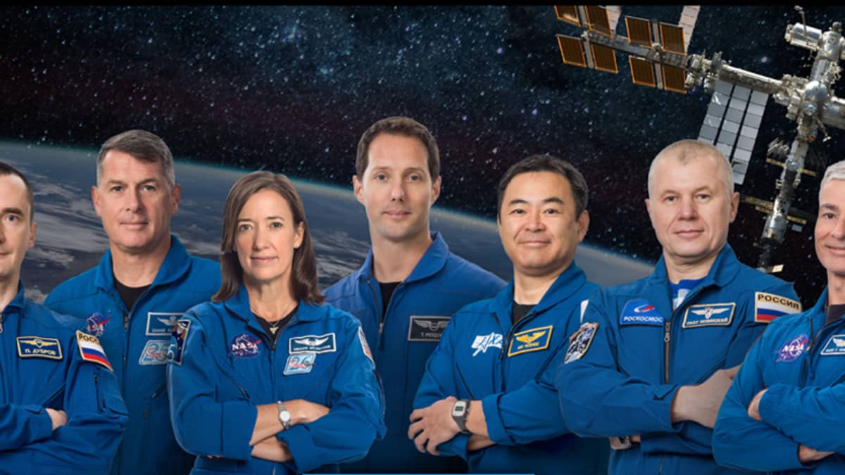 Seitsemän ihmistä avaruslentäjän haalareissa, käsivarret ristissä. Taustalla kuva ISS:stä Maan yllä. 