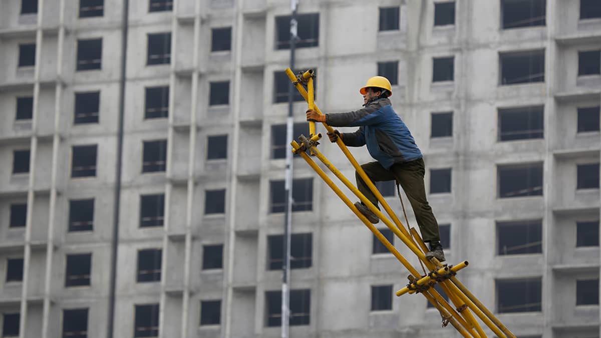 Kiinalainen siirtotyöläinen on kurottautunut kokoamaan metallisia rakennustelineitä Kiinan Pekingissä. Taustalla rakenteilla oleva betoninen kerrostalo.