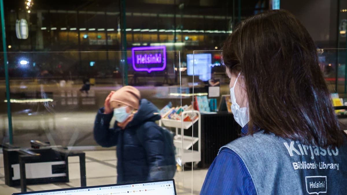 Kirjaston työntekijän näytöllä on teksti "Helmet-lukuhaaste". Työntekijä on näytön edessä selin, selässä teksti "Kirjasto".