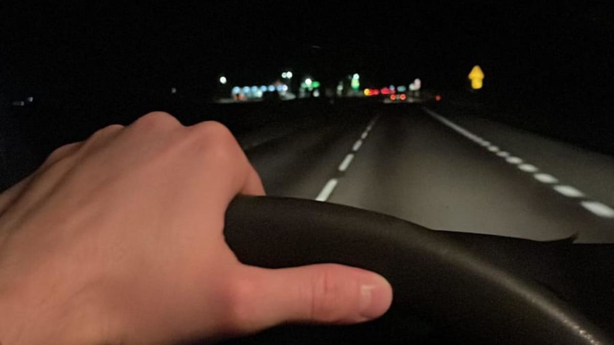 Käsi pitää kiinni auton ratista, edessä näkyy pimeä valtatie.