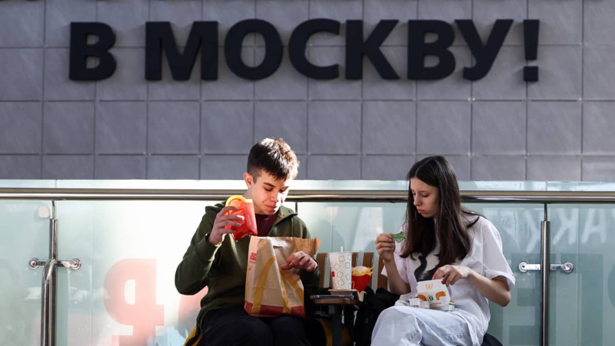 Tyttö ja poika penkillä rautatieaseman edessä syömässä McDonald'sin pikaruokaa. Seinässä lukee venäjäksi "Moskovaan". 