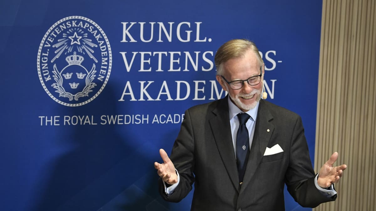 Leväesti hymyilevä Göran Hansson kädet levällään. Taustalla Ruotsin kuninkaallisen tiedeakatemian kyltti sinetteineen. 