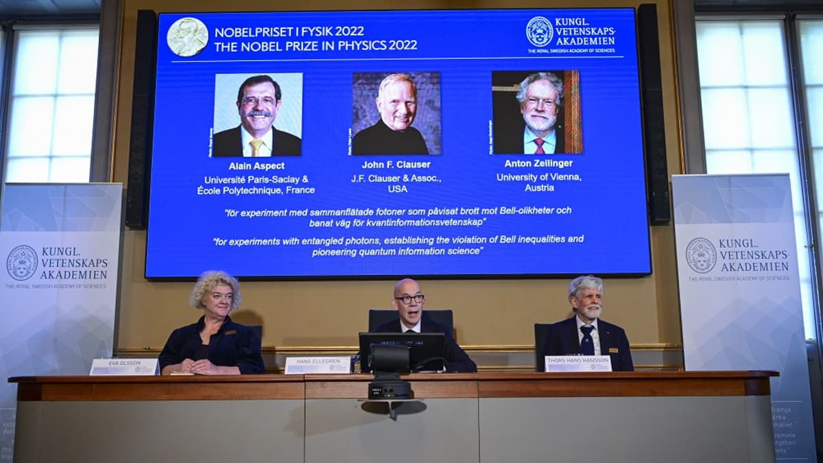 Voittajien kuvat valkokankaalla, edessä kolme Nobel-komitean edustajaa pöydän takana 
