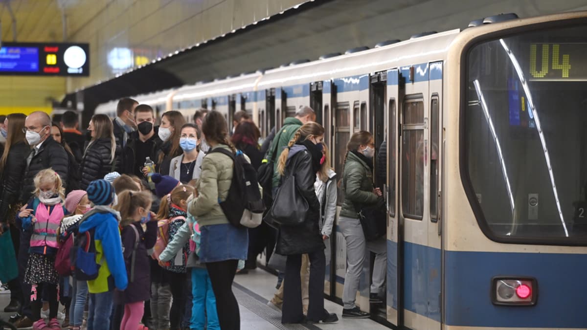 Iso joukko aikuisia ja lapsia nousemassa metrojunaan. Kaikilla on kasvonaamio. 
