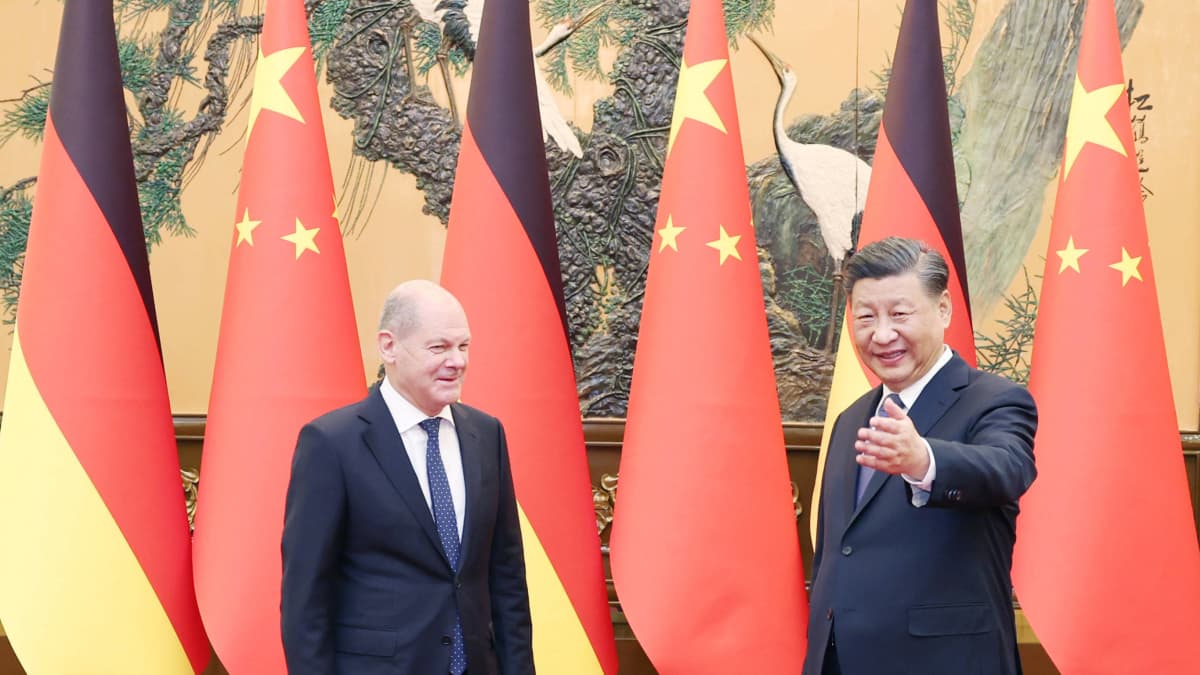 Scolz hymyilee kainosti Xille, joka viittelöi eteenpäin ja katsoo kameraan. Tummapukuiset herrat seisovat maidensa lippujen edessä punaisella matolla. 