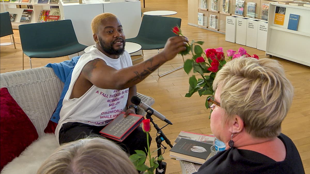Rap-artisti Seksikäs-Suklaa (oik. Luyeye Konssi) ojentaa punaista ruusua rakkausrunoa kuuntelemaan jääneelle rouvalle keskustakirjasto Oodissa 1.8.2019  Puoli seitsemän -ohjelman Lukuhaaste-tempauksessa.