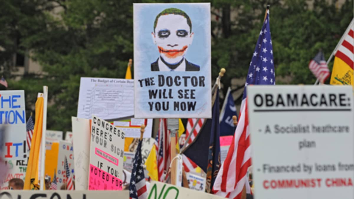 Ihmiset kannattelevat kylttejä Washington DC:n mielenosoituksessa. Keskellä olevassa kyltissä on jokerimainen kuva Barack Obamasta ja teksti 'The Doctor Will See You Now'.