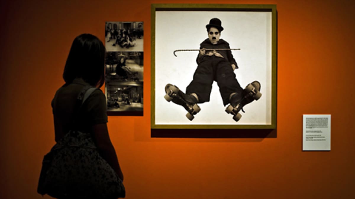 Chaplinin näyttelyssä vieraileva nainen katselee neliömäistä kuvaa Chaplinista punaisella seinällä. Sammakkoperspektiivistä otetussa kuvassa Chaplinilla on knallin ja kävelykepin lisäksi rullaluistimet jaloissaan.