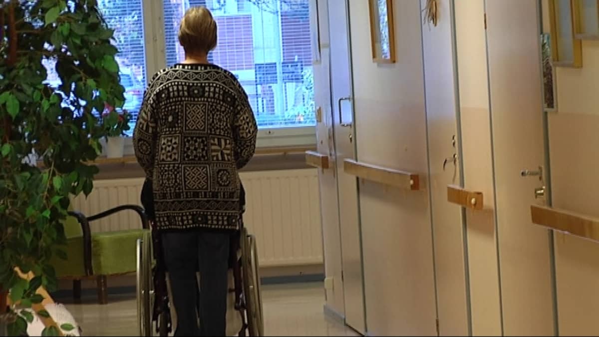 Nainen lykkää vanhusta pyörätuolissa osaston käytävällä.