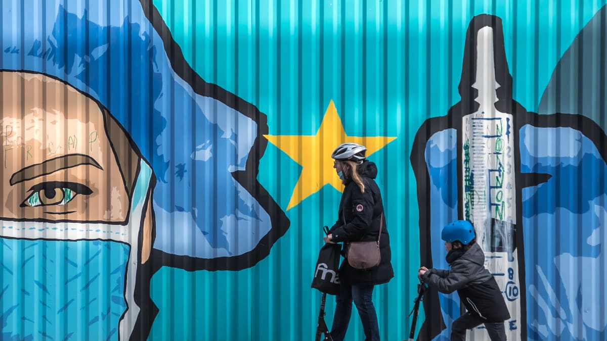 Aikuinen ja lapsi kulkevat potkulaudoilla seinämaalauksen ohi. Maalauksessa lähikuva sairaanhoitajan kasvoista ja rokoteruiskusta.