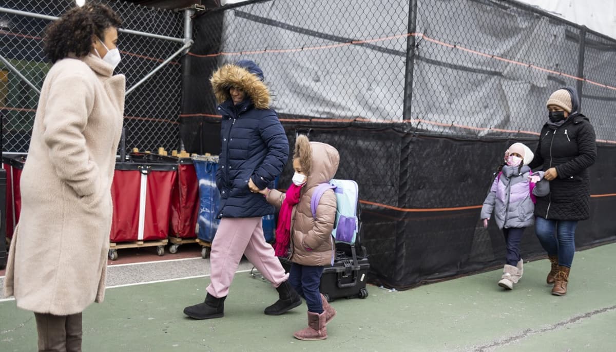 Kuvassa etualalla seisoo nainen. Hänen takanaan kävelee lapsi, jota aikuinen pitää kädestä. Heidän takanaan on toinen lapsi, joka kävelee aikuisen kanssa. 