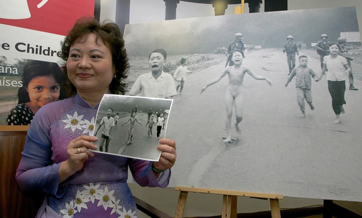 Kim Puc pitää kädessään kuvaa itsestään ja muista juoksevista lapsista, joiden takana kävelee Yhdysvaltain sotilaita. Taustalla on suurennos kuvasta.