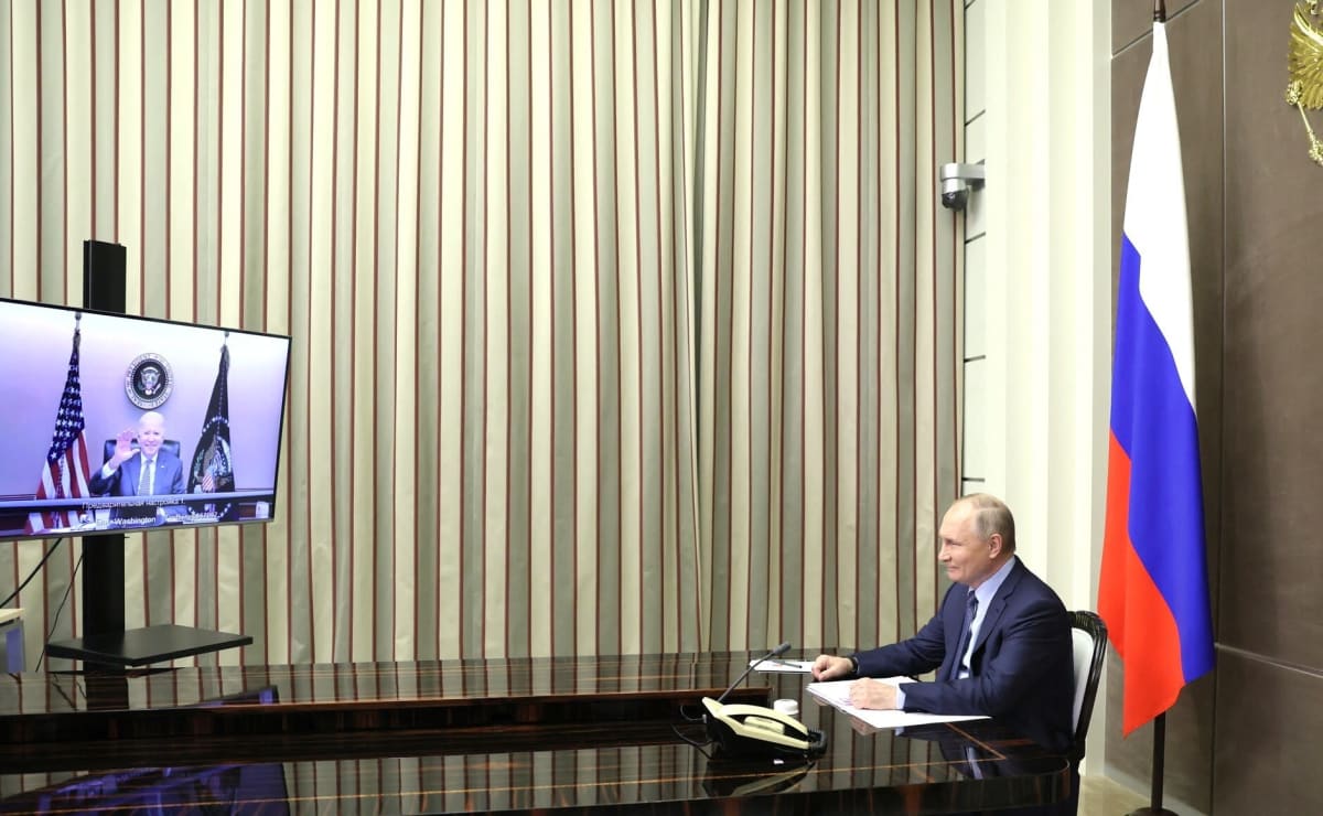 Vladimir Putin istuu myhäillen kuvan oikeassa laidassa ja katsoo monitoria, josta Joe Biden vilkuttaa. 