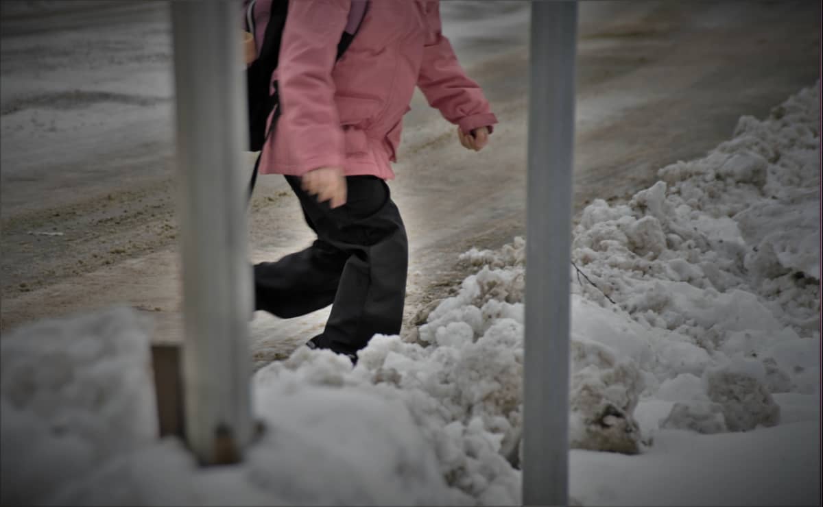 Koululainen ylittää lumisen tienreunan. Suojatie on huonosti aurattu lumisateen jälkeen.