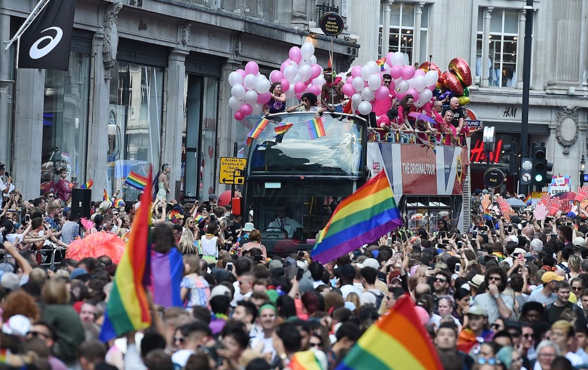 Katu on täynnä marssijoita, ja monet kantavat sateenkaaren värisiä lippuja. Kuvan keskiosassa keskellä ihmismerta on kaksikerroksinen bussi, jonka avoimessa yläosassa on ihmisiä valkoisten ja vaaleanpunaisten ilmapallojen kanssa.