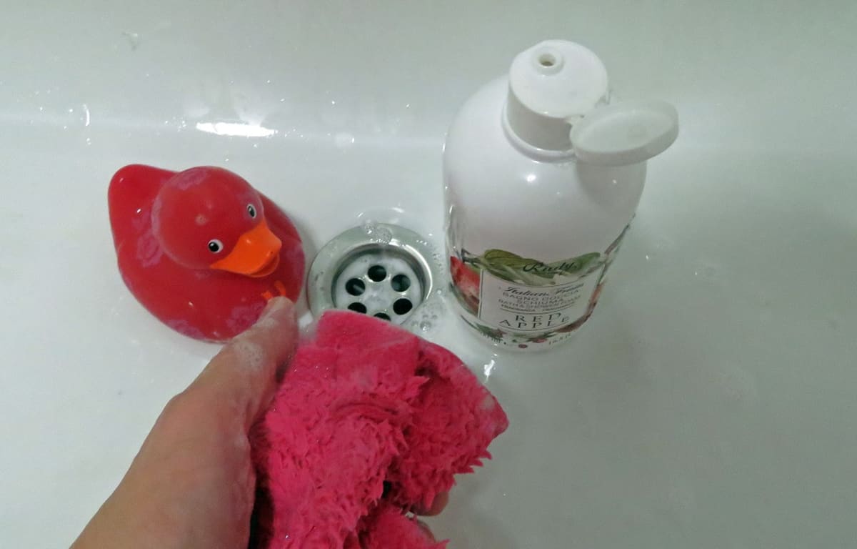 Kylpyankka ja suihkuvaahtopullo kylpyammeen pohjalla. Kädessä pesulappu, jossa saippuavaahtoa.