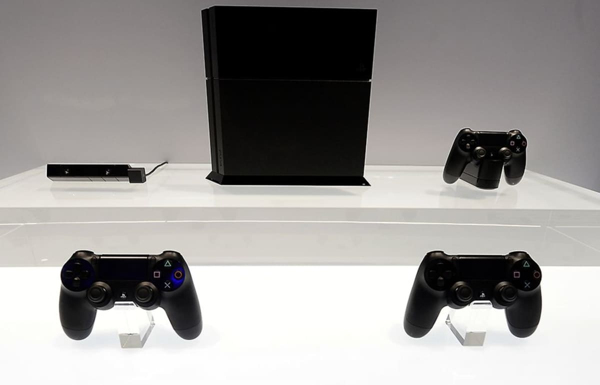 Väriltään musta playstation 4 -pelikonsoli ja  mustia peliohjaimia esillä E3-messuilla kirkkaan valkoista alustaa vasten.